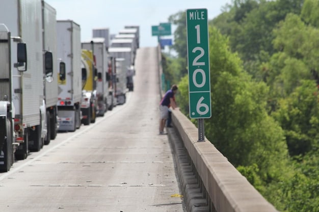 A long line of semi-trucks backed up at a US-Mexico border crossing point, with customs officers visible, indicating potential delays due to new tariff compliance or increased checks.